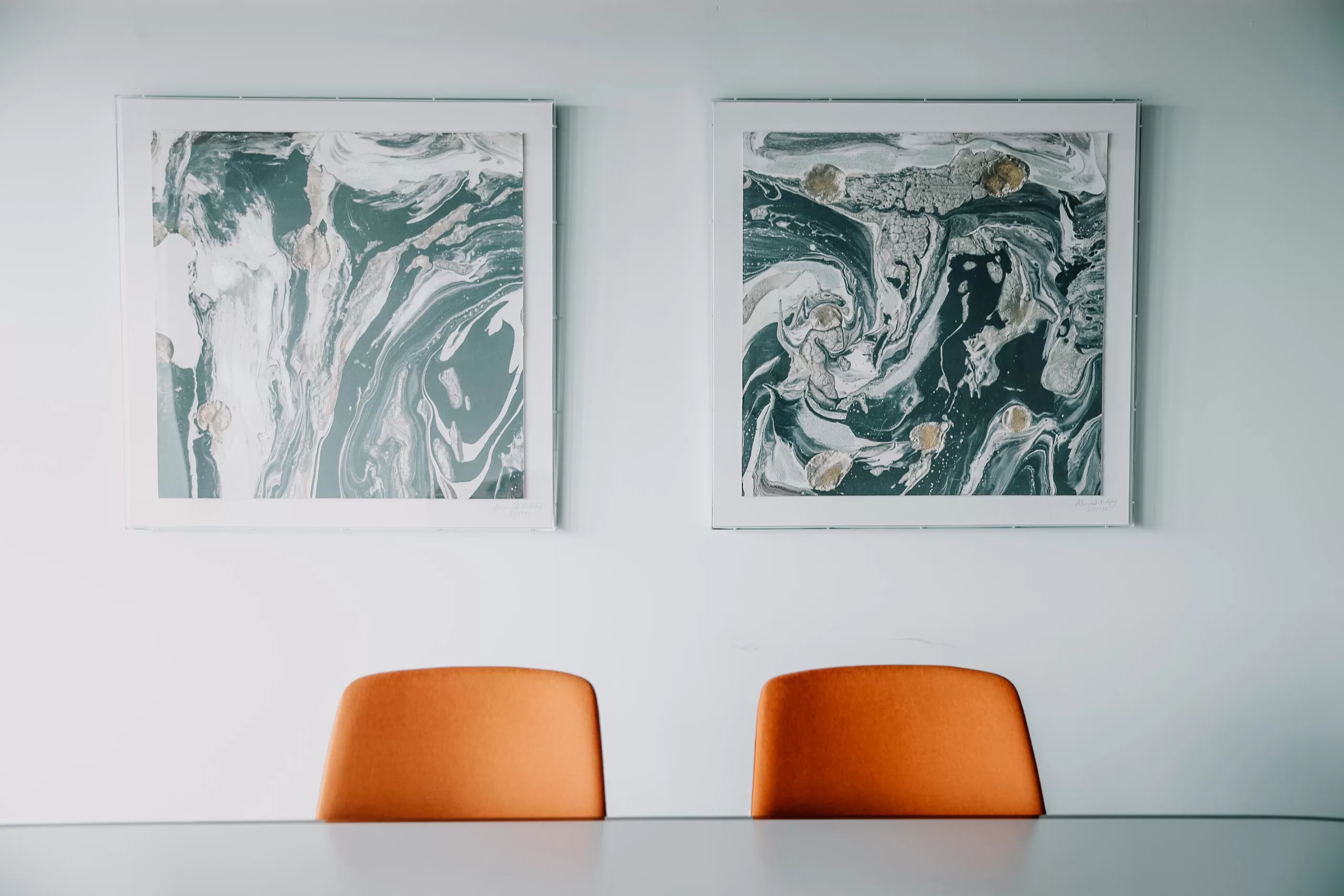 Two abstract paintings above orange chairs in a white room.