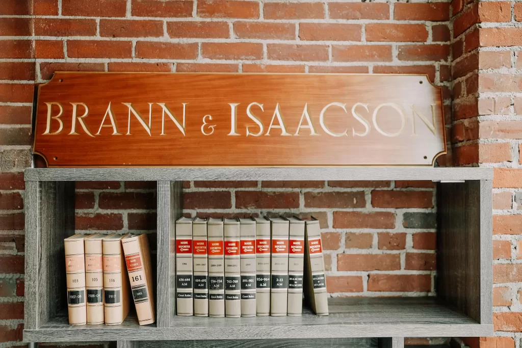 Wooden sign reading "Brann & Isaacson" above law books on a shelf, brick wall background.